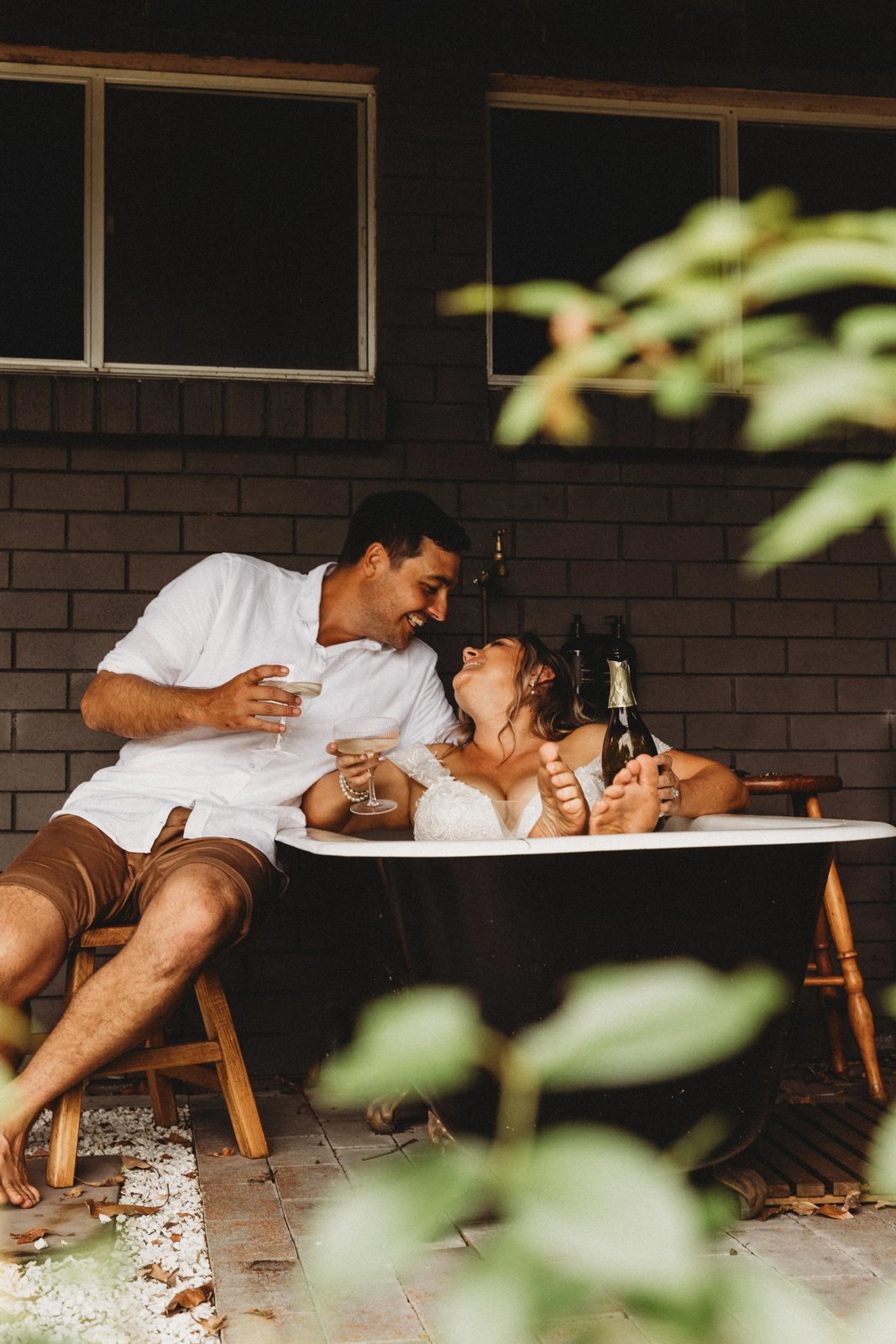 A couple laughing in an outdoor bathtub with champagne, the bride in white