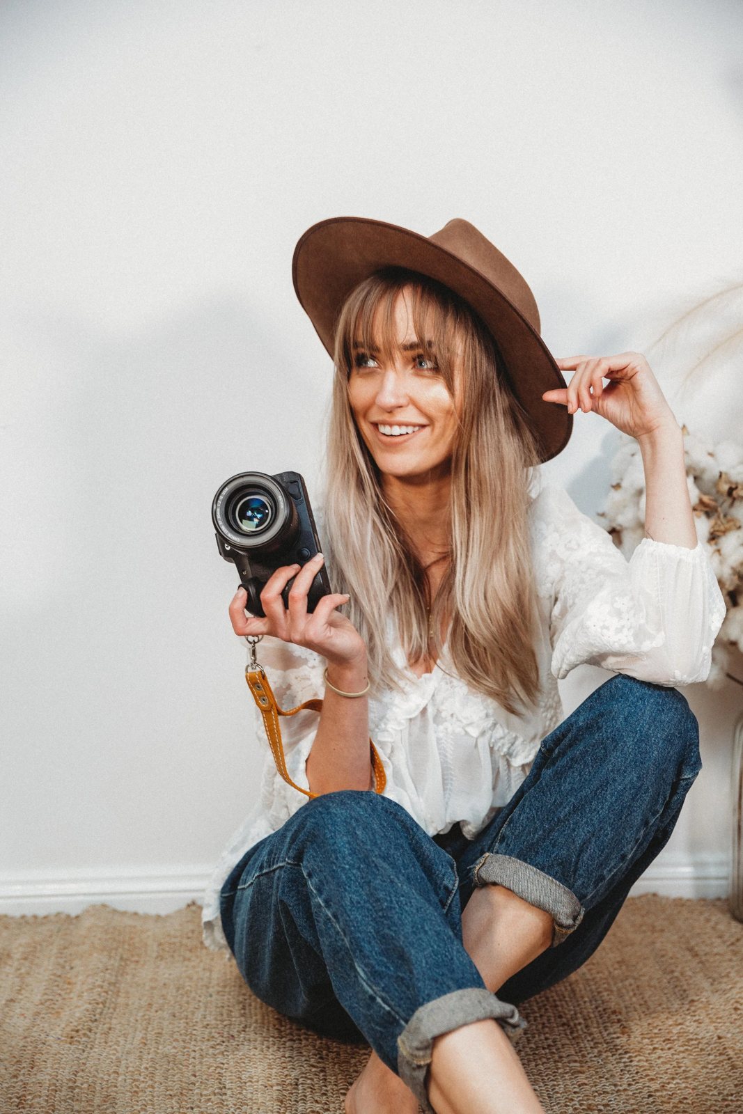 Bec Slade sitting on the floor in a brown hat and jeans, smiling, with her camera in hand
