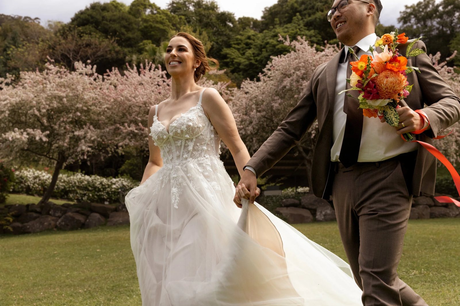 A bride and groom walking hand in hand past pink blossom trees, an orange bouquet in his hand
