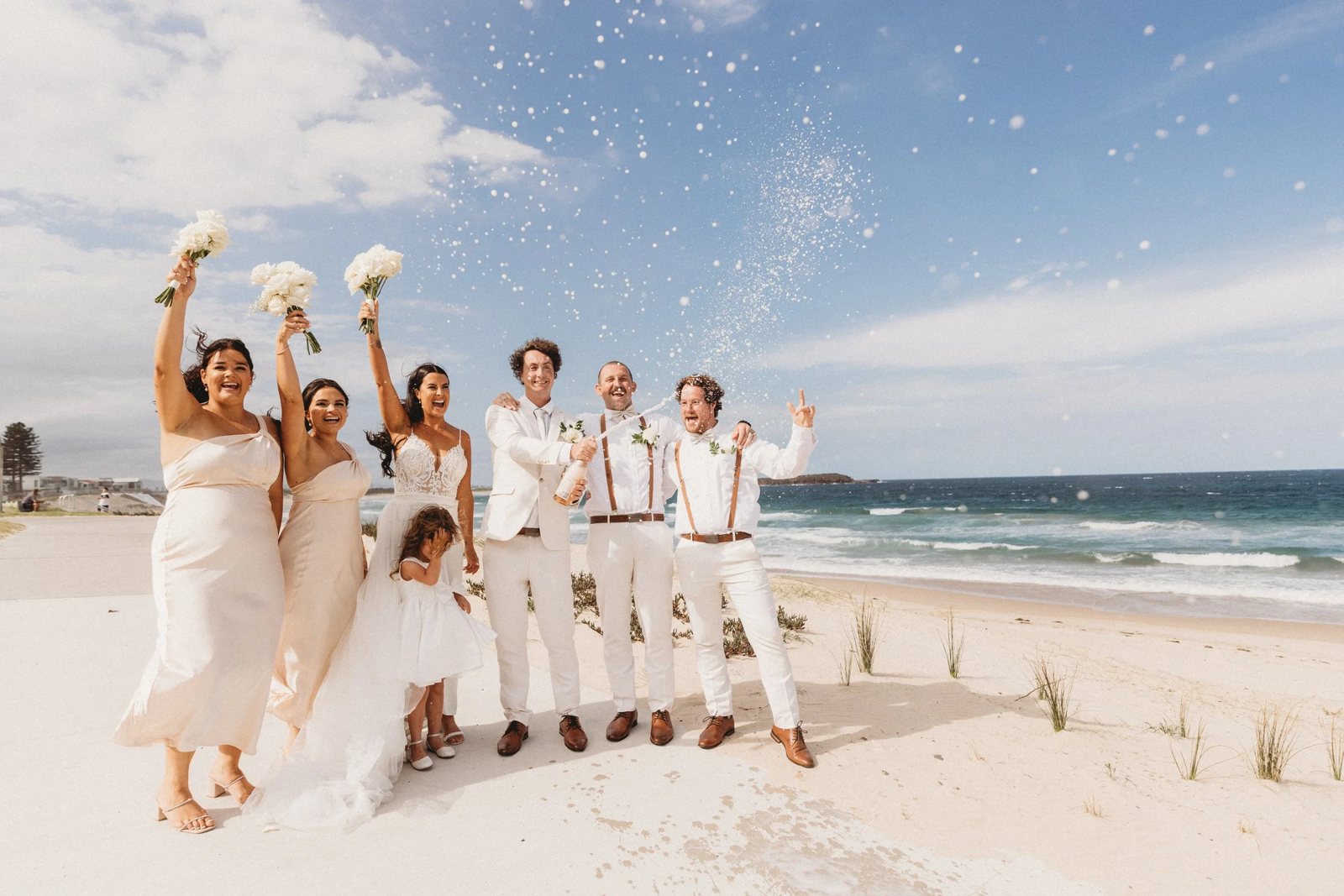 A bridal party in white jumping on the beach with bouquets in the air