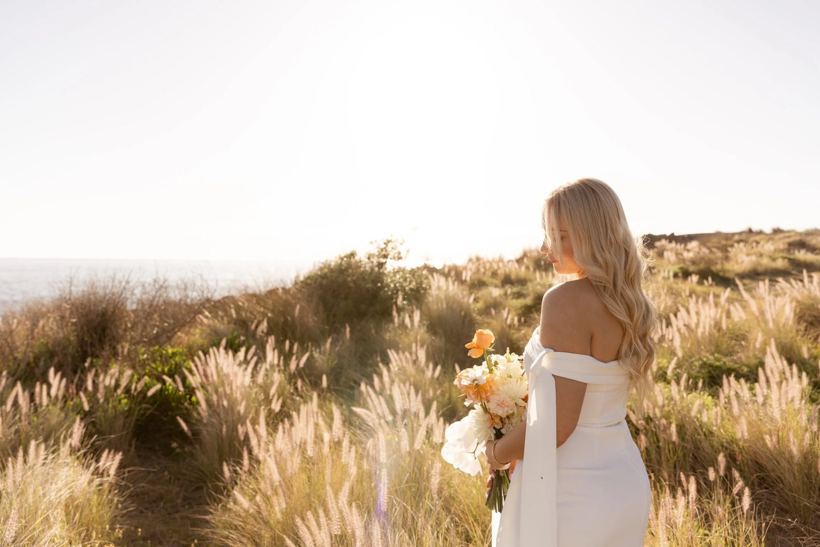 A bride with a peach bouquet standing in coastal grass, backlit by the sun