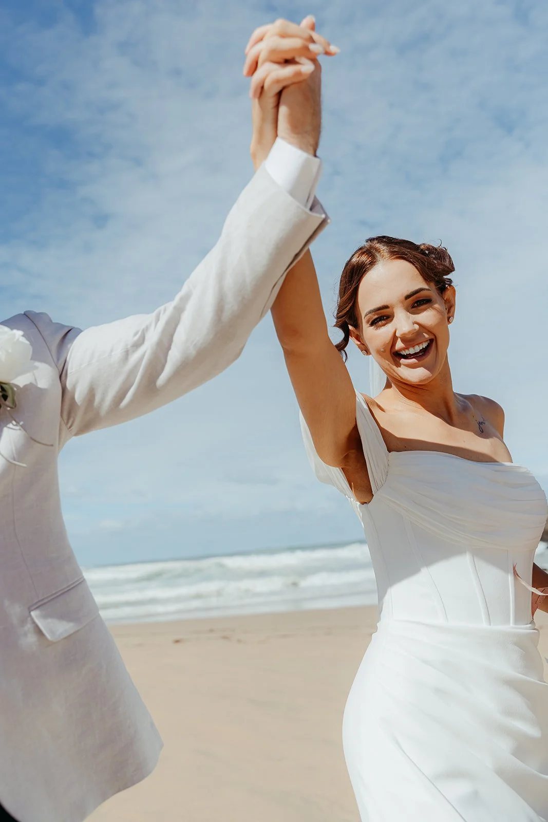 A bride laughing on the beach, her hand raised high in her partner&rsquo;s