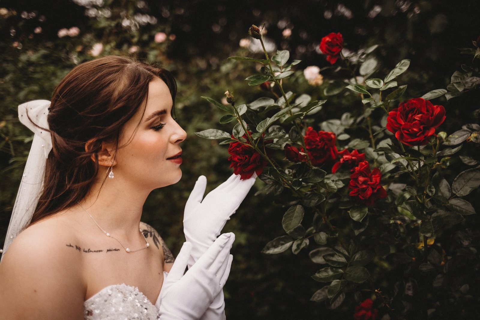 A bride in white gloves leaning towards a bush of red roses