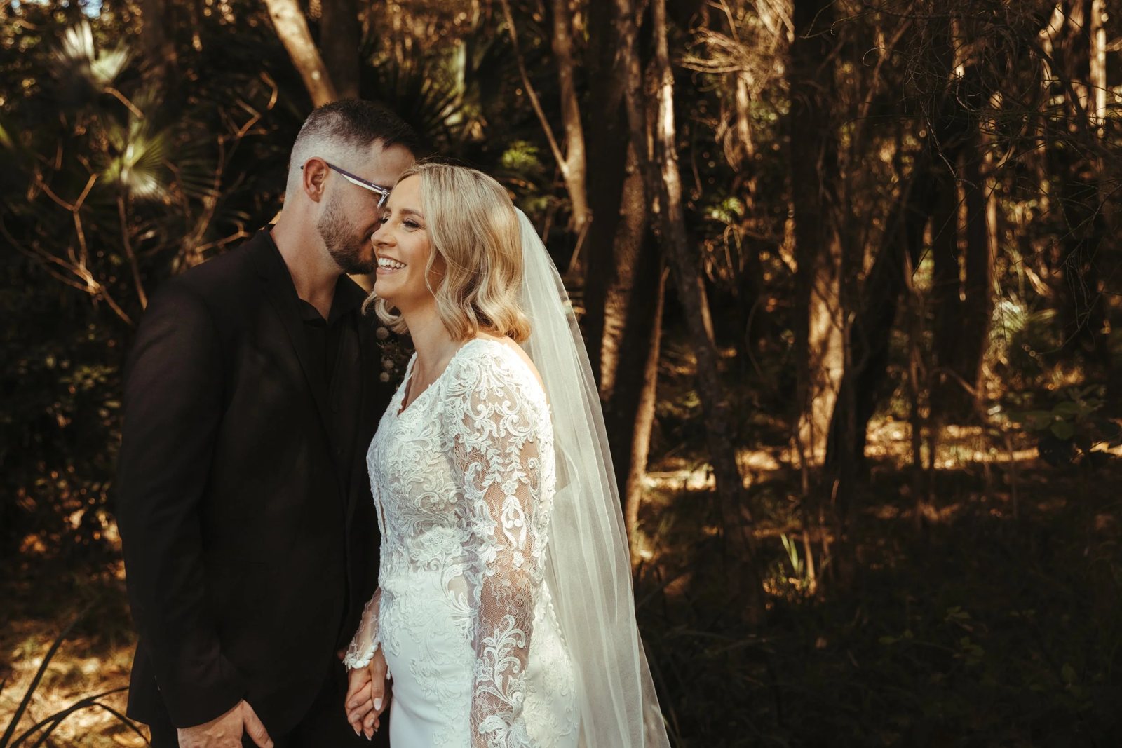 A bride in a lace gown and veil and her groom in black, foreheads close in a dark forest
