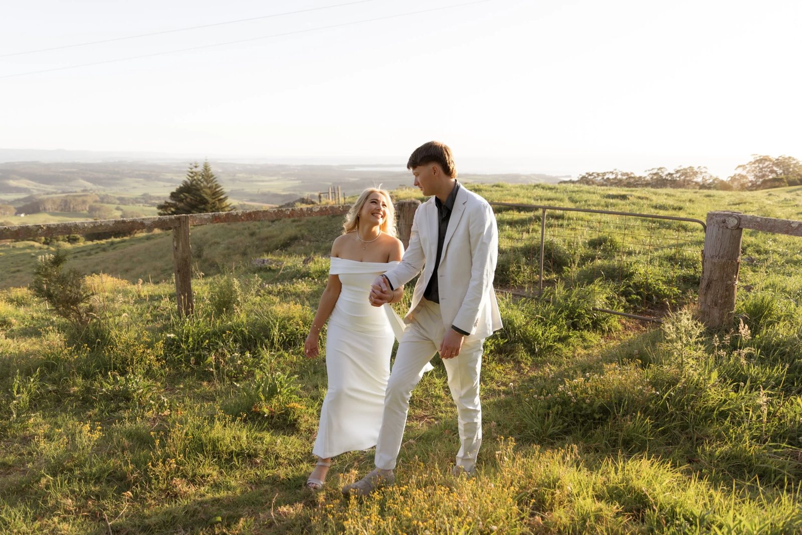 A bride and groom walking hand in hand across a green Kiama hillside at golden hour