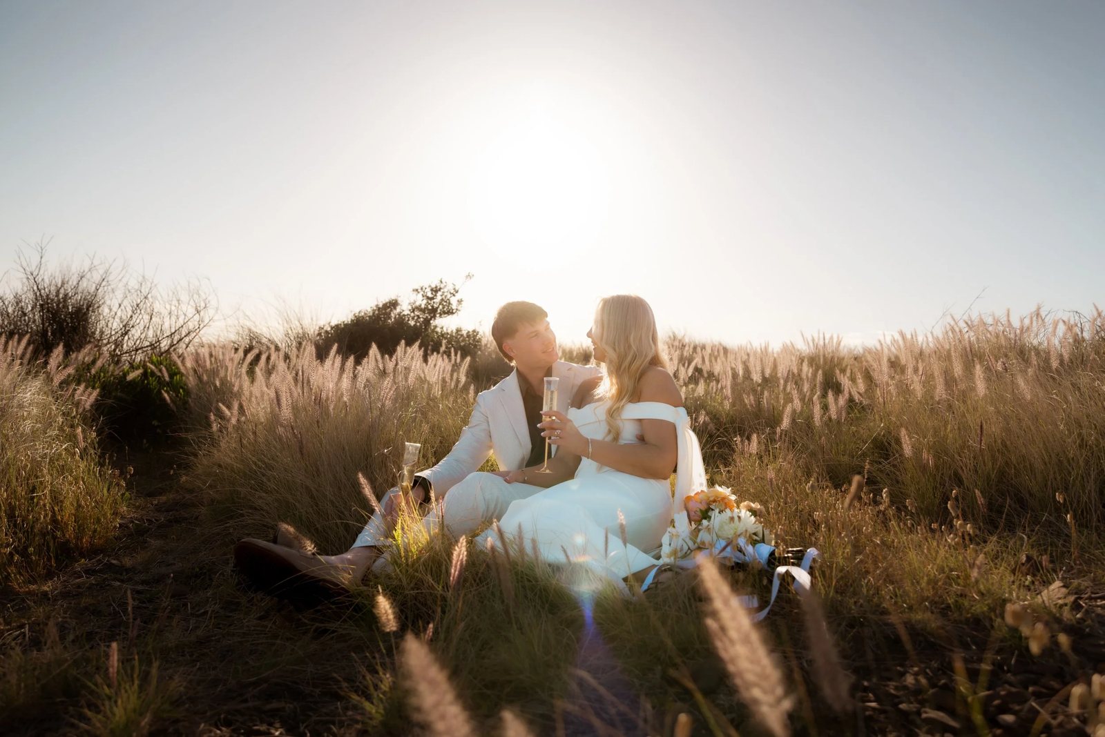A couple in wedding clothes sitting in tall golden grass at sunset, glasses raised