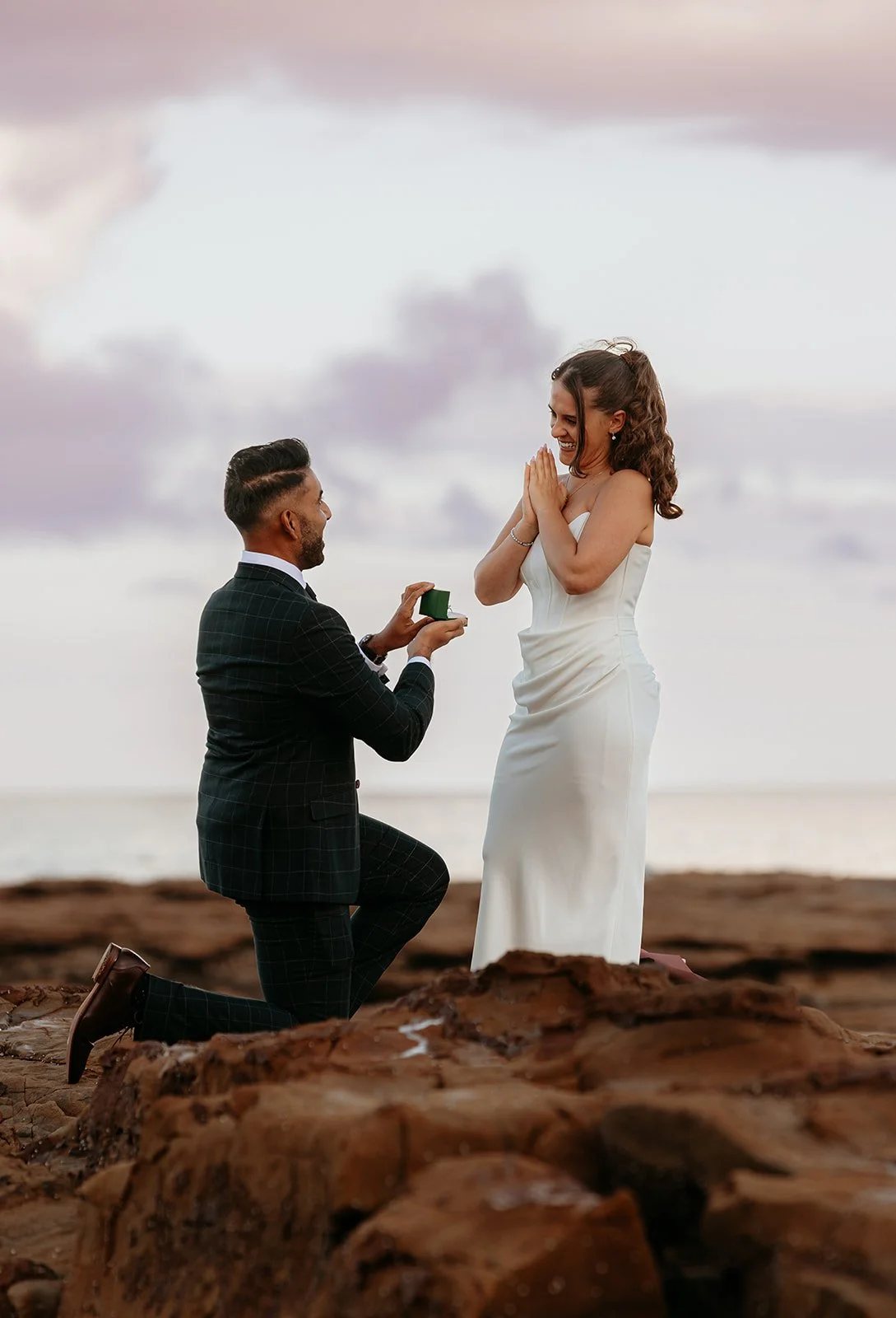 A man on one knee holding out a ring box to a woman on the rocks at sunset, her hands to her face