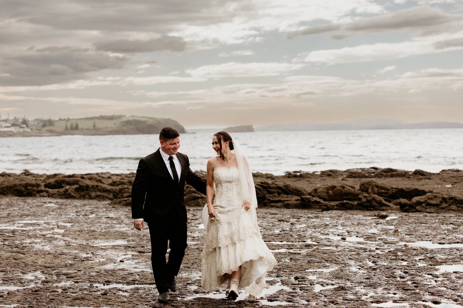A bride and groom walking hand in hand across a rock shelf by the sea