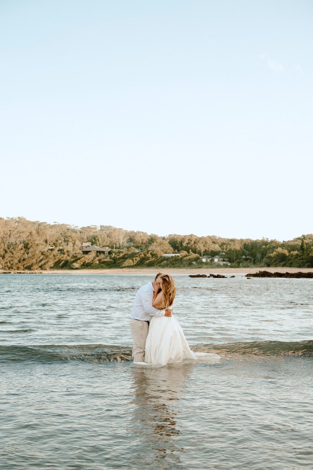 A couple kissing waist-deep in a calm bay, the bride&rsquo;s dress floating