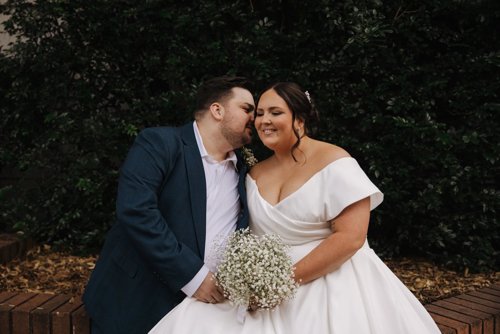 A groom kissing his bride on the cheek as she holds a bouquet of baby&rsquo;s breath